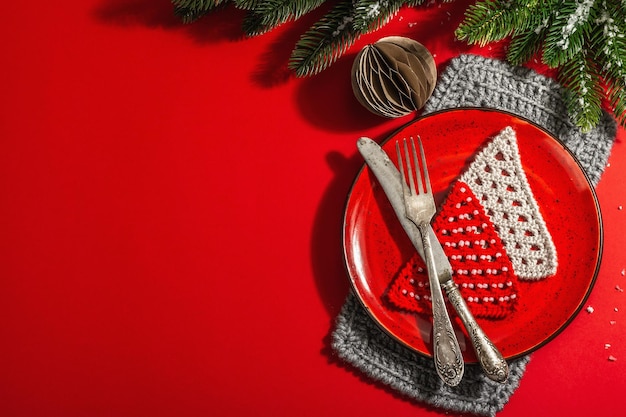 Serviert weihnachtliche Tischdekoration in Rottönen mit Vintage-Besteck. Immergrüne Äste, gehäkelte Tannen, Kunstschnee. Modernes hartes Licht, dunkler Schatten. Flache Lage, roter Hintergrund, Draufsicht