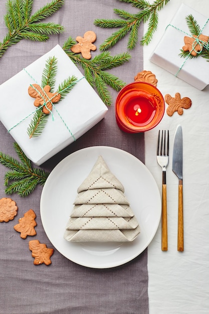 Servilleta en forma de árbol de Navidad en un plato sobre un mantel blanco con regalos y decoraciones con ramitas de abeto y galletas de jengibre