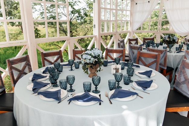 Servindo e decoração da mesa redonda de casamento na varanda ao ar livre Placas de óculos azuis e pratos em uma mesa festiva com flores