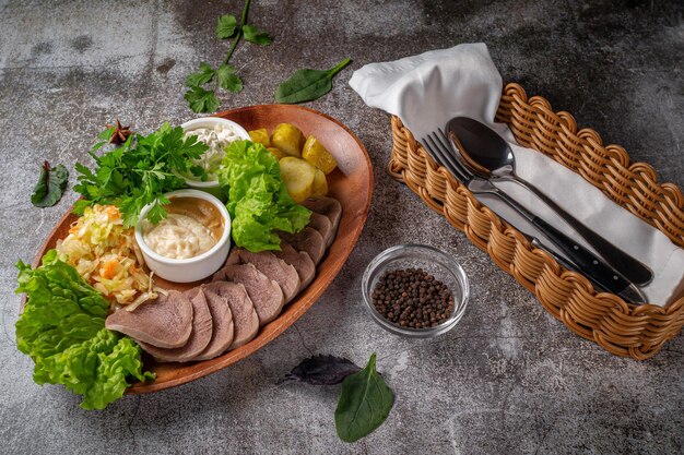 Servindo um prato do menu do restaurante. Língua de boi fatiada com picles, repolho, molhos e ervas em um prato contra uma mesa de pedra cinza, um delicioso aperitivo