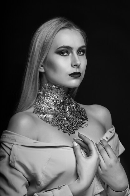 Sesión de belleza en blanco y negro: impresionante modelo rubia posando en el estudio con maquillaje brillante y papel de aluminio en el cuello