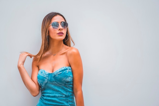 Sesión de estilo de vida, una joven morena caucásica con un vestido de mezclilla azul y gafas de sol sobre un fondo gris liso, con espacio para copiar y pegar, mirando hacia la derecha tocando su cabello