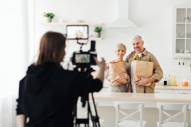 Sesión de estudio y familia en casa con compras de comestibles. Fotógrafo con cámara hace fotos. Un anciano alegre y una dama adulta sostienen bolsas ecológicas de papel con compras en el interior de la cocina minimalista