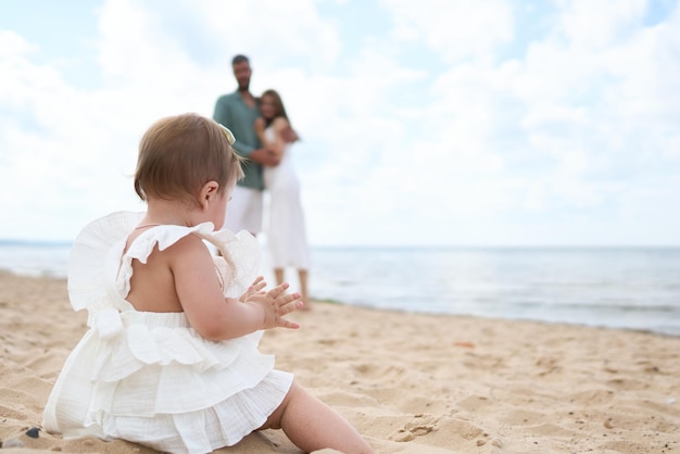 Una sesión de fotos de padres felices admirando a su niña de un año jugando con arena en la playa