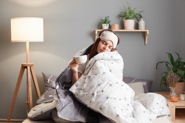 Sesión interior de una mujer somnolienta con los ojos vendados sentada en la cama envuelta en una manta sosteniendo una taza de café tomando una siesta posando con los ojos cerrados durmiendo