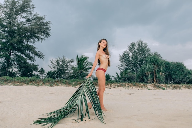 Sessão de fotos tropical na areia. Jovem bonita elegante posa em pé, cobrindo o peito com a mão e segurando a folha de palmeira. Um olhar para o lado. Fundo de praia com árvores exóticas e céu.