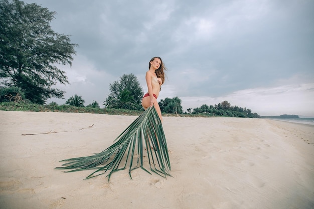 Sessão de fotos tropical na areia. Uma jovem esbelta linda posa de pé na praia nua cobrindo o peito com a mão e segurando uma folha de palmeira olhando para longe.