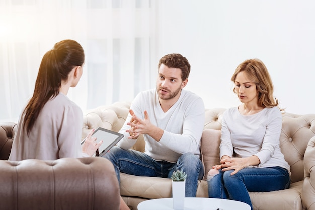 Sessão psicológica. Bonito homem barbudo infeliz sentado com sua esposa no sofá e olhando para o psicólogo enquanto explica o problema dela para ela