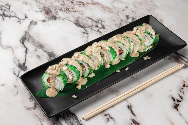 Set aus Maki-Rollen mit Chuka-Wakame-Lachs-Nori-Sesam-Sauce mit Nüssen und grünem Bambusblatt in einer schwarzen Keramikplatte mit Essstäbchen auf einem hellweiß strukturierten Marmorhintergrund, Seitenansicht