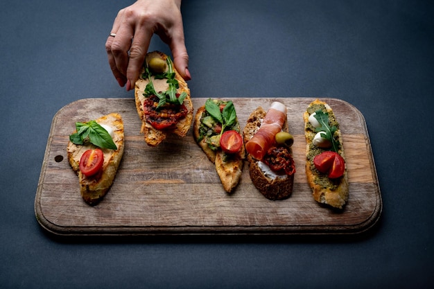 Set Bruschettas mit Jamon, Oliven, Pesto, Guacamole, Tomaten, Basilikum, serviert auf Holzbrett. Die Hand der Person nimmt traditionelles mediterranes geröstetes Brot mit Rucola und Gemüse