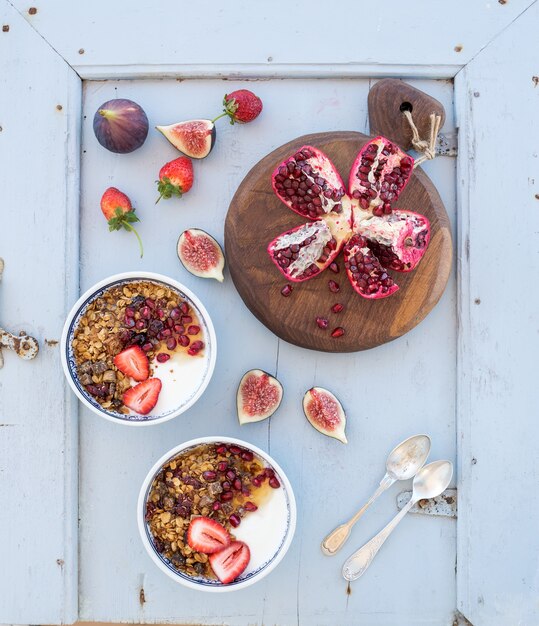 Set de desayuno saludable. Tazones de granola de avena con yogurt, fresas frescas, higos, granada y miel sobre una mesa de madera azul claro