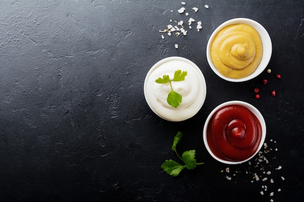 Set mit drei Saucen - Mayonnaise, Senf und Ketchup in weißen Keramikschalen auf schwarzem Stein oder Beton.