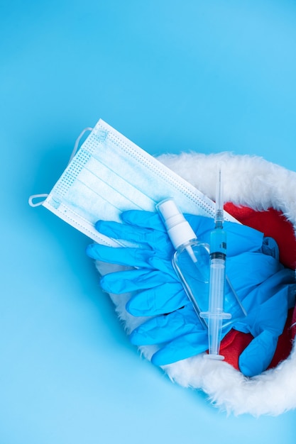 Set de regalo para las vacaciones de Navidad y Año Nuevo 2021. Un conjunto higiénico de mascarilla médica, guantes, un desinfectante y una jeringa con una vacuna y un chip de coronavirus en un gorro de Papá Noel. Vacunación 2021.