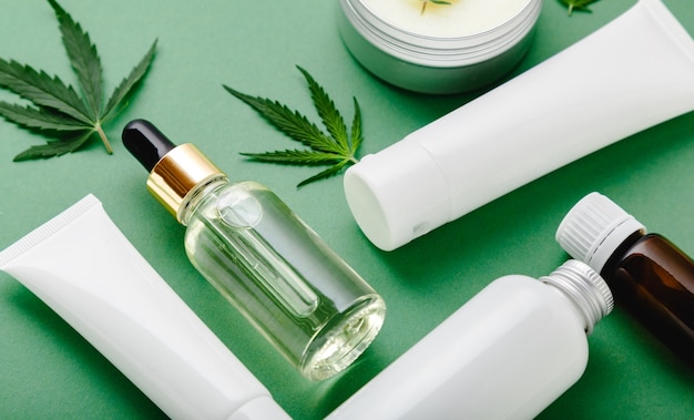 Set von Hanf-Hautpflegekosmetik in weißer Mockup-Verpackung. Feuchtigkeitscreme, Serum, Lotion, CBD-Öl, ätherisches Öl aus Cannabisblättern. Flach auf grünem Hintergrund legen.