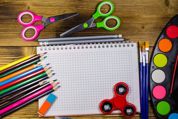 Foto set von schulpapieren. leeres notizbuch, buntstifte, stifte, aquarellfarben, scheren, pinsel, radiergummi und zappelspinner auf holzschreibtisch. zurück zum schulkonzept