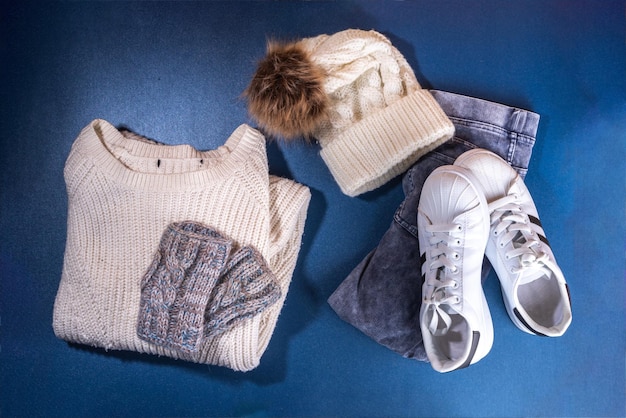 Set weiblicher Winter-Herbst-Kleidung, Jeans, weißer Strickpullover und Mütze, Fäustlinge und Turnschuhe auf dunkelblauem Hintergrund, lässiges, gemütliches Mode-Outfit, flach liegend