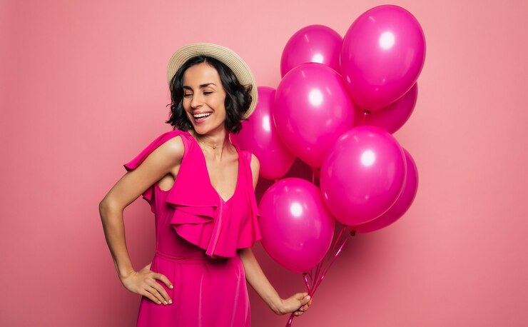 Foto seu aniversário perfeito. jovem mulher encantadora em um vestido fúcsia e chapéu de palha está segurando balões rosa na mão esquerda e rindo.
