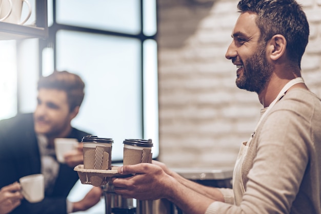 Seu café para viagem! Vista lateral do barista passando xícaras de café para seu cliente com um sorriso no balcão do bar