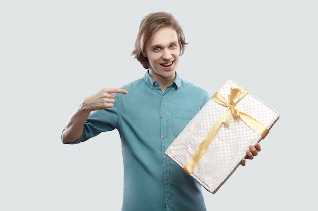 É seu! Homem jovem moderno satisfeito na camisa azul clara em pé e apontando o dedo para apresentar com laço amarelo e sorriso cheio de dentes, olhando para a câmera. Interior, isolado, foto de estúdio, fundo cinza