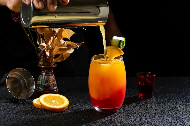 SEXO DE CÓCTEL EN LA PLAYA, HOMBRE DE BAR PREPARANDO UN CÓCTEL, PONIENDO ZUMO DE NARANJA EN LA BEBIDA, EN FONDO OSCURO