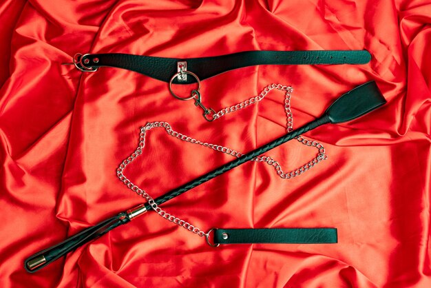 Sexspiele für Erwachsene BDSM-Artikel Lederriemenhalsband mit Metallkettenleine und Peitsche auf einem roten Satinblatt