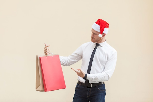 Sexta-feira negra no inverno. empresário de cabeça vermelha de felicidade com chapéu de papai noel, segurando sacolas de compras nas mãos, apontando o dedo e olhando para presentes. foto interna, fundo laranja claro