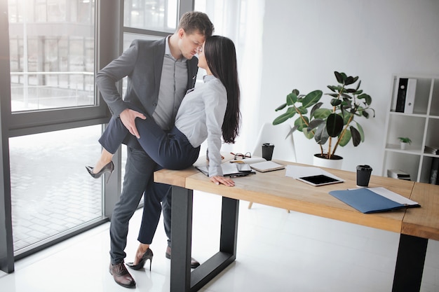 Sexuelles und intimes Bild eines Paares bei der Arbeit. Sie sitzt auf dem Tisch. Er hält ihr Bein in sexueller Pose.