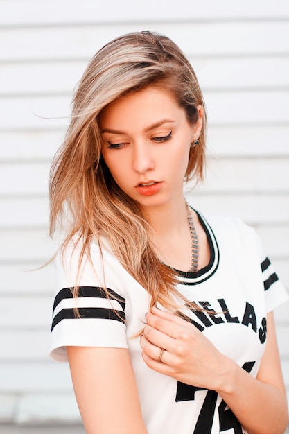 Sexy amerikanisches Mädchen mit blonden Haaren in einem modischen weißen T-Shirt mit einem schwarzen Muster posiert in der Nähe einer weißen hölzernen Vintage Wand. Modernes attraktives Mädchen. Lebensstil.