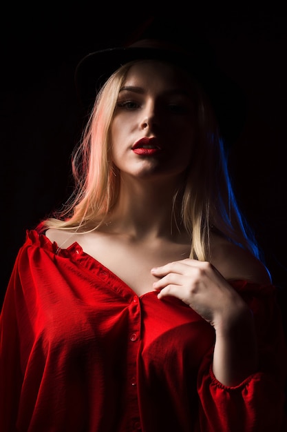 Sexy blonde Frau mit roter Bluse und Hut posiert im Schatten mit blauem und rotem Licht