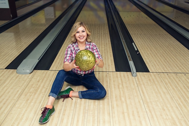 Sexy blondes Mädchen, das mit Ball auf Boden im Bowlingclub sitzt.