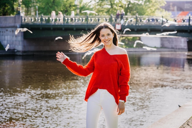 Sexy brünette junge schöne Frau in rotem Pullover und weißer Hose geht am Fluss spazieren und wirft lose Haare