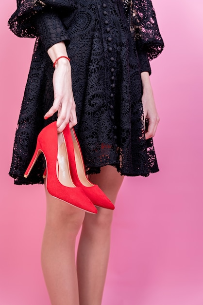 Sexy Brunettefrau im schwarzen Kleid, das rote Schuhe auf rosa Hintergrund hält