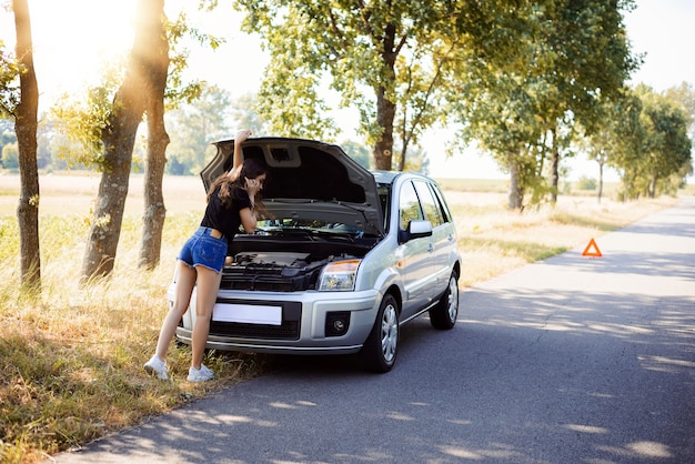 Sexy chica morena con piernas largas desnudas de pie cerca de coche roto