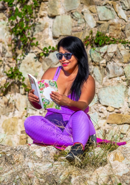 Sexy, dunkelhäutige, lateinamerikanische Frau mit rosa, engen Leggings und Top, die draußen ein Buch liest