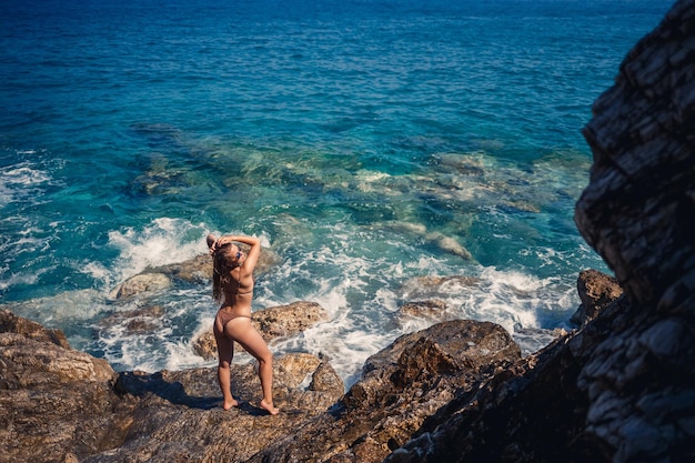 Sexy Frau im Ganzkörper-Badeanzug mit langen Haaren geht auf großen Felsen an einem felsigen Strand während eines Sturms auf See Rückansicht Frau Bademode Insel Tropen Landschaft exotischer Spaziergang