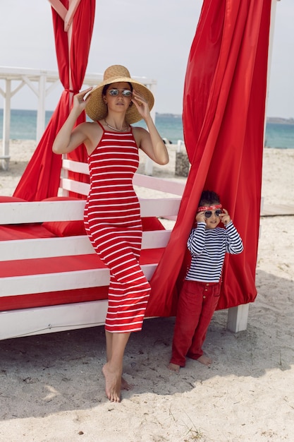 Sexy Frau in einem rot gestreiften Kleid und einem Strohhut steht im Sommer mit einem Baby in der Nähe einer überdachten Sonnenliege am Strand