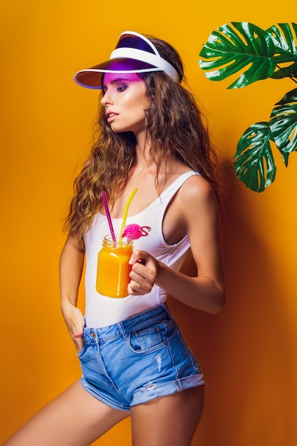 Sexy Frau in weißen Badeanzug und Blue Jeans Shorts, trendiges Visier hält Glas frisches Getränk, während auf gelb, orange heiß stehen