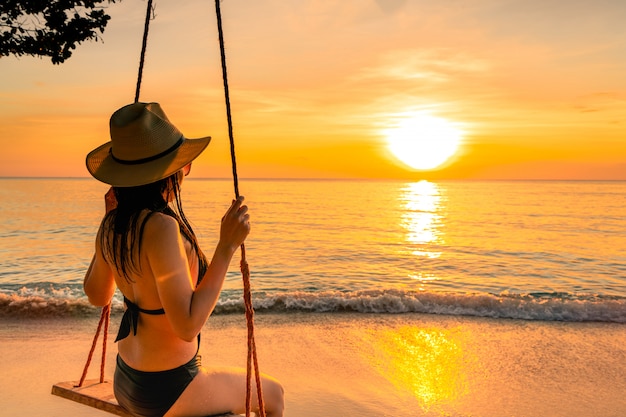 Sexy Frau tragen Bikini und Strohhut schwingen die Schaukeln am tropischen Strand in den Sommerferien bei Sonnenuntergang.