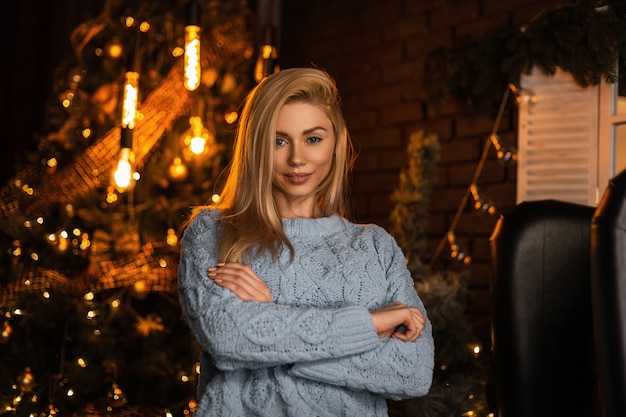 Sexy glückliches junges Modell einer schönen blonden Frau in einem stilvollen Strickpullover, der in einem dunklen Raum gegen einen hellen festlichen Weihnachtsbaum mit Weinlesegirlanden aufwirft