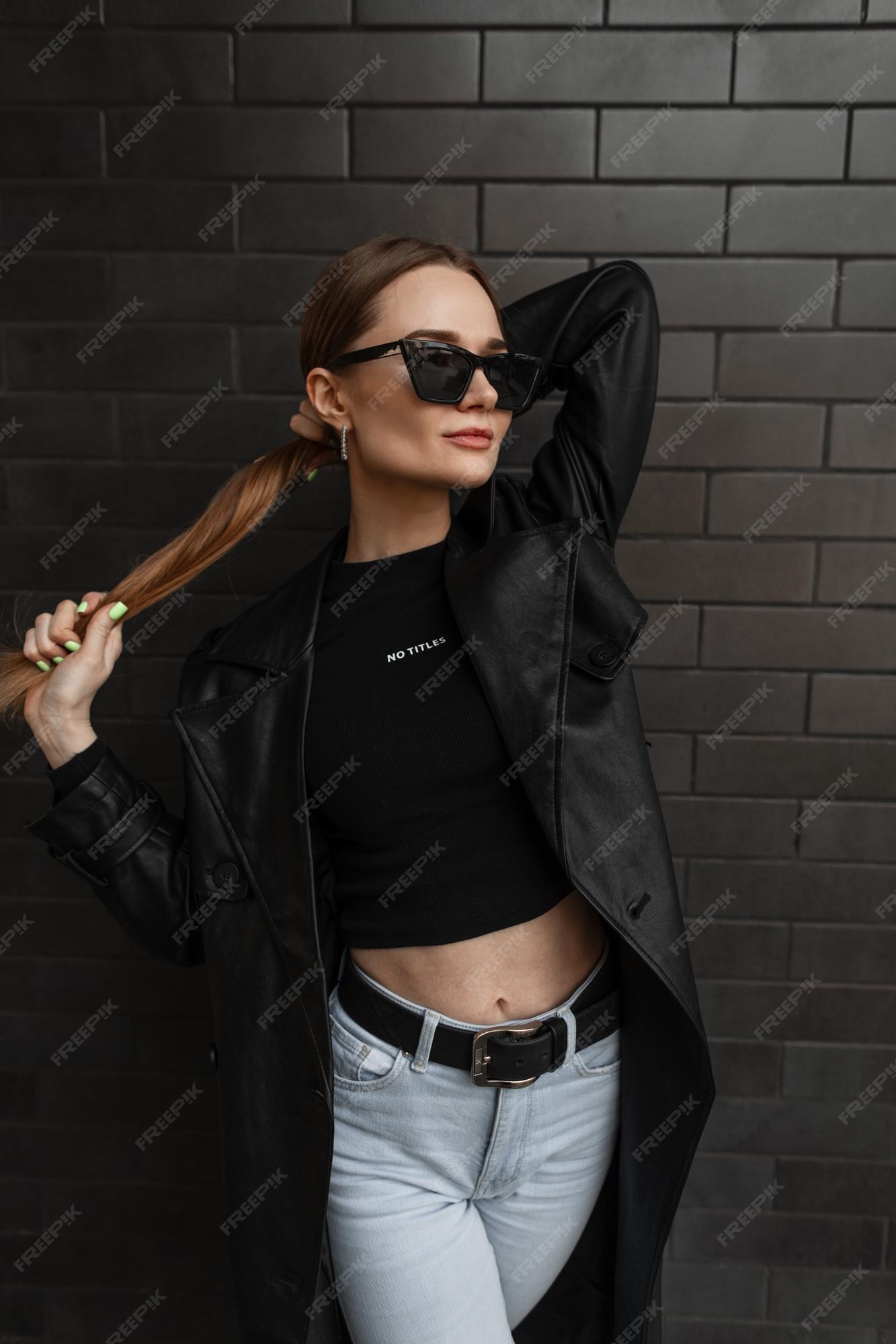 Sexy hermosa modelo de mujer de moda en elegante ropa de calle negra con una chaqueta larga de cuero negro camiseta jeans y de sol de pie cerca de una pared