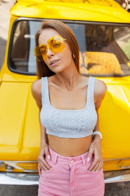 Sexy hermosa mujer brillante con elegantes gafas de sol amarillas en un top de verano de moda y jeans rosas de moda se encuentra junto a un auto amarillo retro vintage en una vista superior de día soleado Vacaciones de verano y positividad