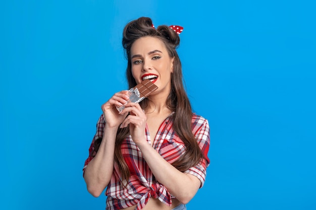 Sexy jovem pinup com roupa de estilo retrô comendo barra de chocolate no fundo azul do estúdio