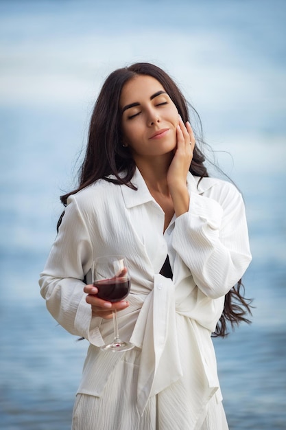 Sexy junge Brünette in einem schwarzen Bikini und weißem Anzug mit einem Glas Wein posiert am Strand