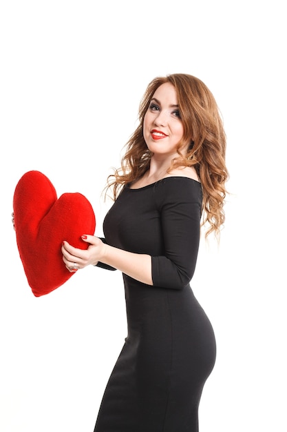 Sexy junge Frau des Valentinstags mit den roten Lippen, die das selbst gestaltete Herz im schwarzen Kleid halten. Modefrau, die schwarzes Kleid mit großem rotem Herzen trägt. Valentinstag.