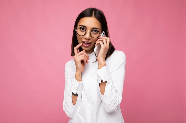 Sexy linda jovem morena vestindo blusa branca e óculos ópticos em pé isolado sobre um fundo rosa, falando no celular, olhando para a câmera e pensando.