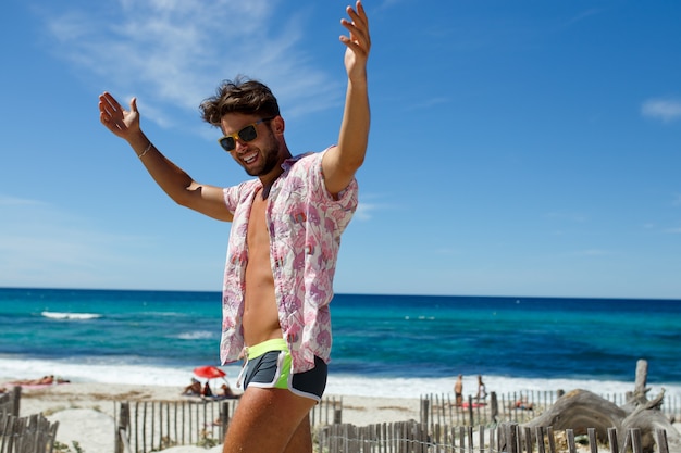 Sexy Modell des Athleten in der Badebekleidung und in der Sonnenbrille, die auf dem Strand mit den auf Korsika-Insel, Frankreich erhobenen Armen aufwirft. Urlaub, Sommer, Meer, Himmel Hintergrund. Horizontale Ansicht.