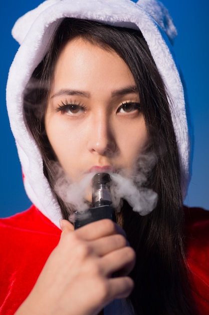 Sexy mujer china en traje de santa claus fumando cigarrillo electrónico y exhalando vapor