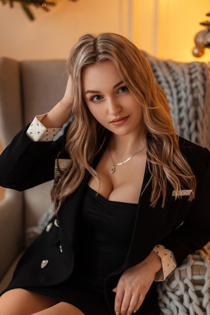 Sexy mujer hermosa con una cara linda en un elegante traje negro de moda y una chaqueta de lujo con exuberantes pechos se sienta y descansa en un sillón en el interior