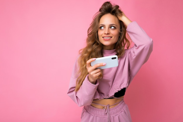 Sexy mujer joven rubia atractiva con sudadera con capucha rosa aislada sobre fondo rosa con espacio de copia sosteniendo y usando teléfono móvil mirando hacia el lado.