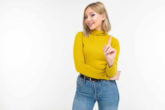 Sexy mujer joven rubia en jeans y blusa apretada amenaza con el dedo índice sobre un fondo blanco.